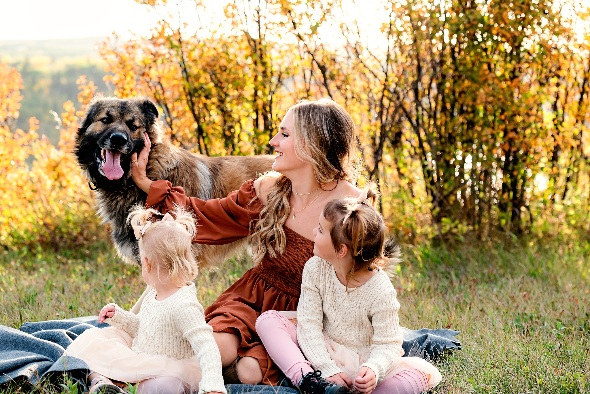 motherhood photography session mom and daughters sit together near autumn colored shrubs farm dog trying to photobomb outdoor fall family photography session lacombe county ab