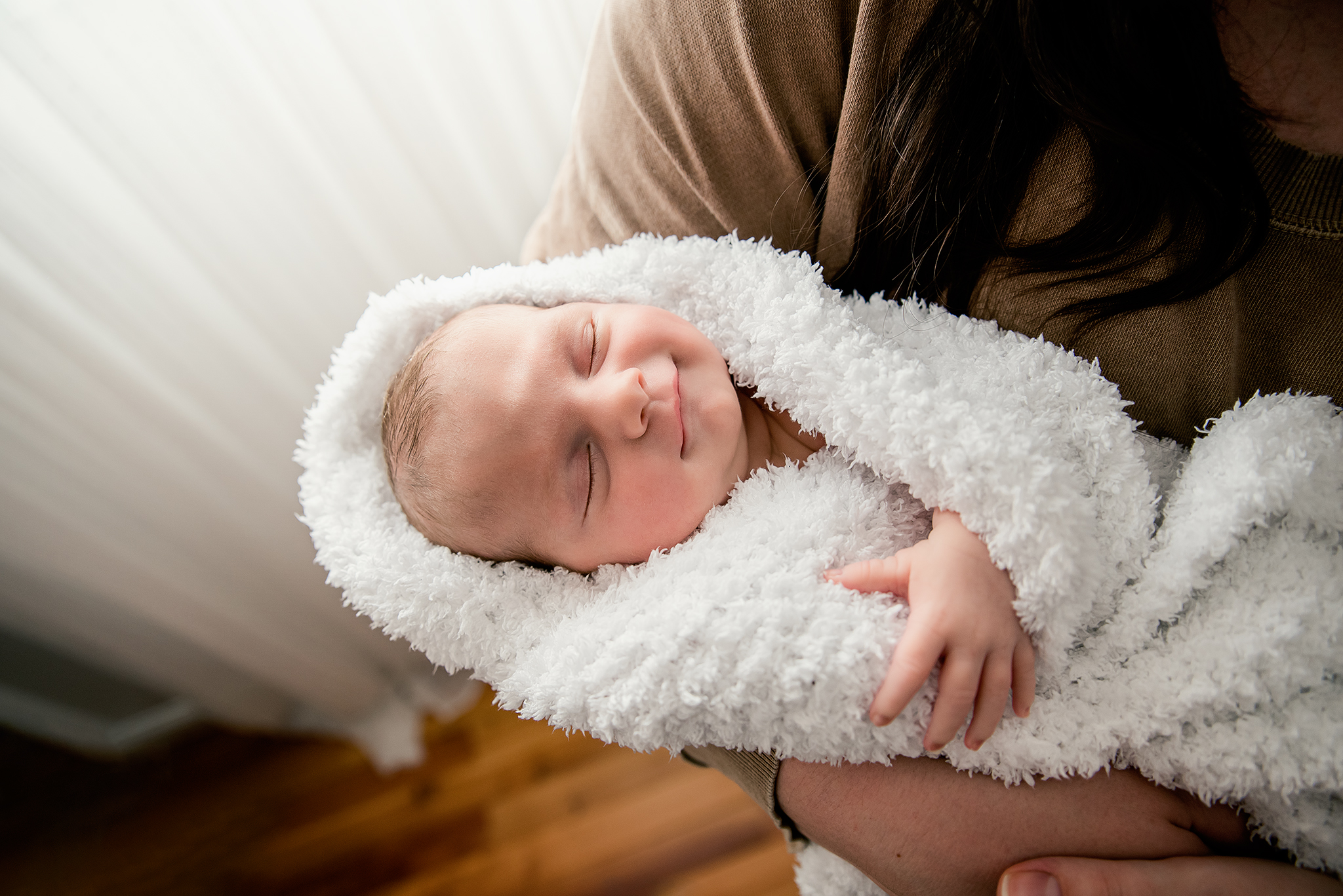 newborn baby girl sleeping in moms arms smiling wrapped in hand made knit fuzzy blanket white curtains in home lifestyle photo session edmonton ab