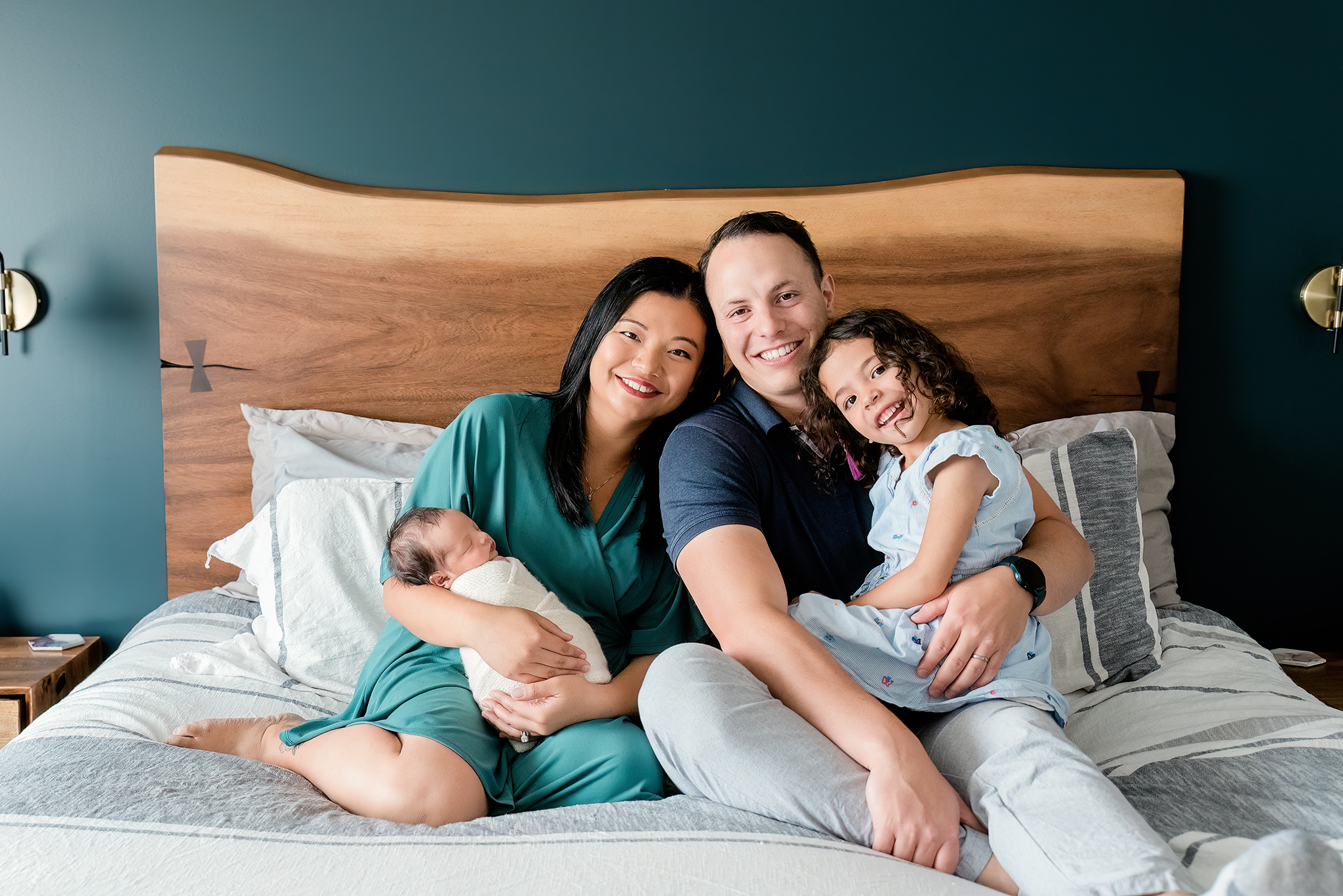 family of four sitting on master bed walnut live edge headboard feature smiling at camera in home newborn photography sherwood park ab