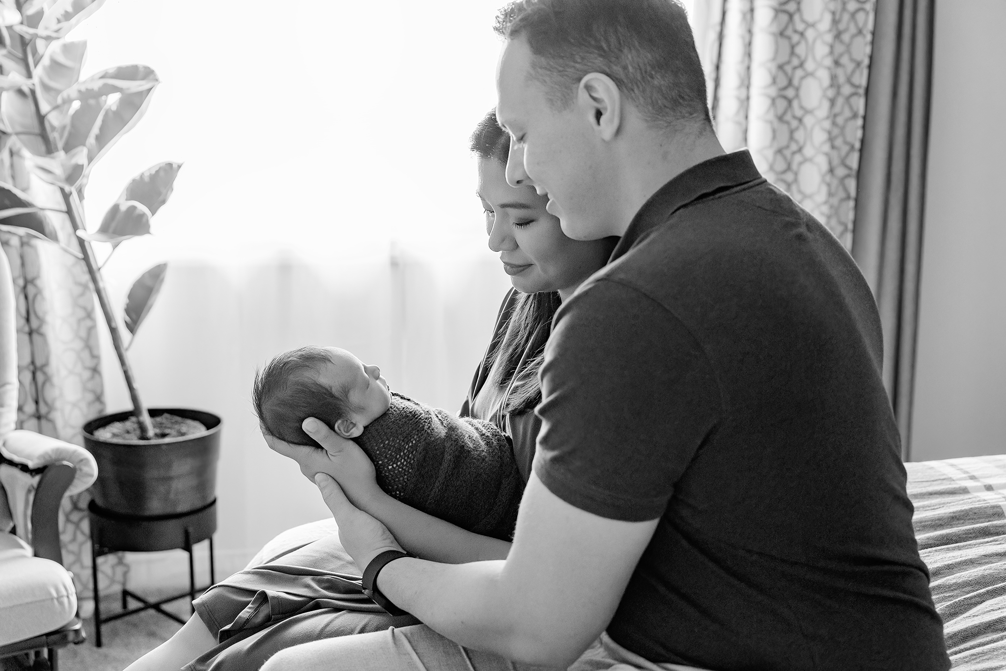 mom and dad sit on bed holding baby boy smiling down at him back lit through sheer curtains in home newborn session bw