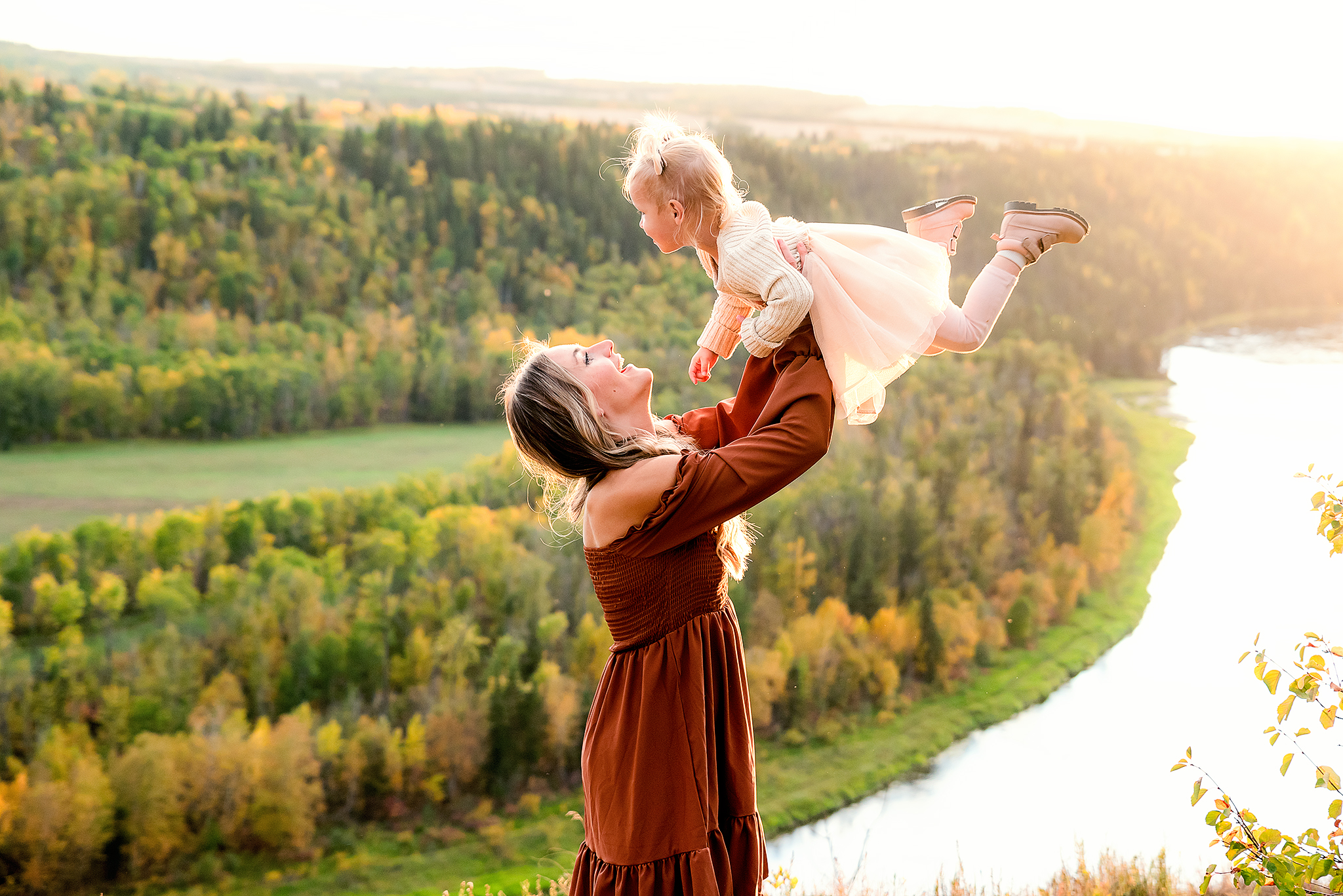 mom plays with young daughter holding her up in the air scenic river valley beneath them golden hour motherhood fall photo session